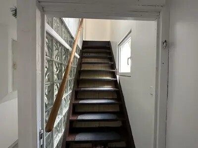 Schmale Innentreppe aus dunklen Holzstufen mit Geländer im hellen Eingangsbereich, links Glasbausteinwand.
