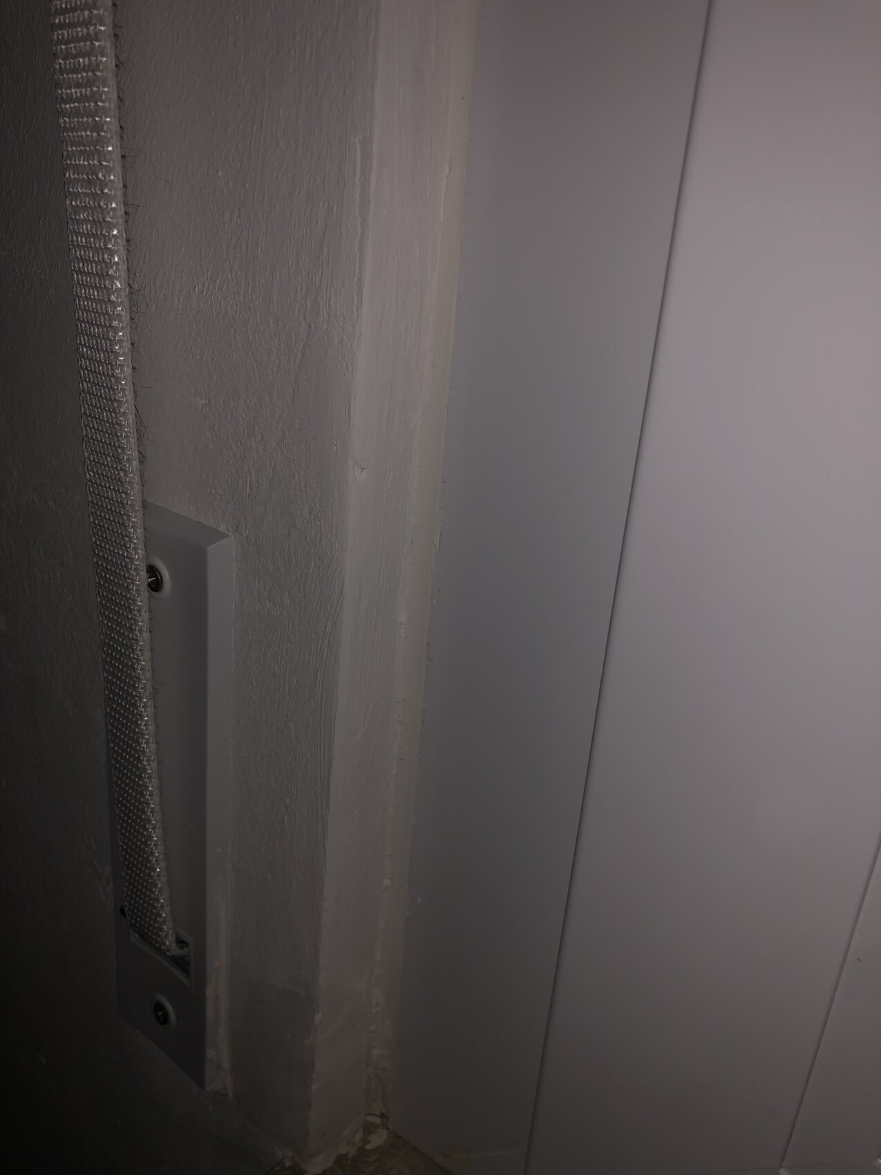 Nahaufnahme einer grauen Wandkante: links eine vertikale Metallleiste neben einer glatten Türverkleidung rechts.