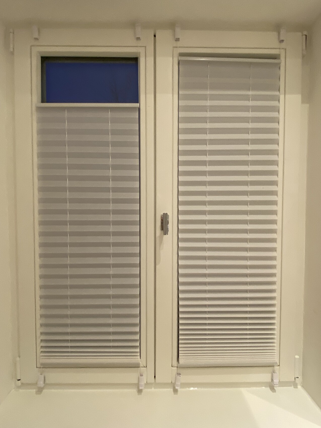 Zwei weiße Fenster mit horizontalen Jalousien; linkes Fenster hat Oberlicht mit blauem Himmel.
