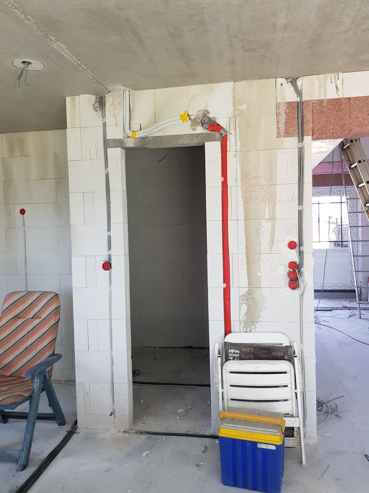 Innenraum einer Baustelle mit weißen Fliesen, rotem Installationsrohr, Stühlen und Kiste.