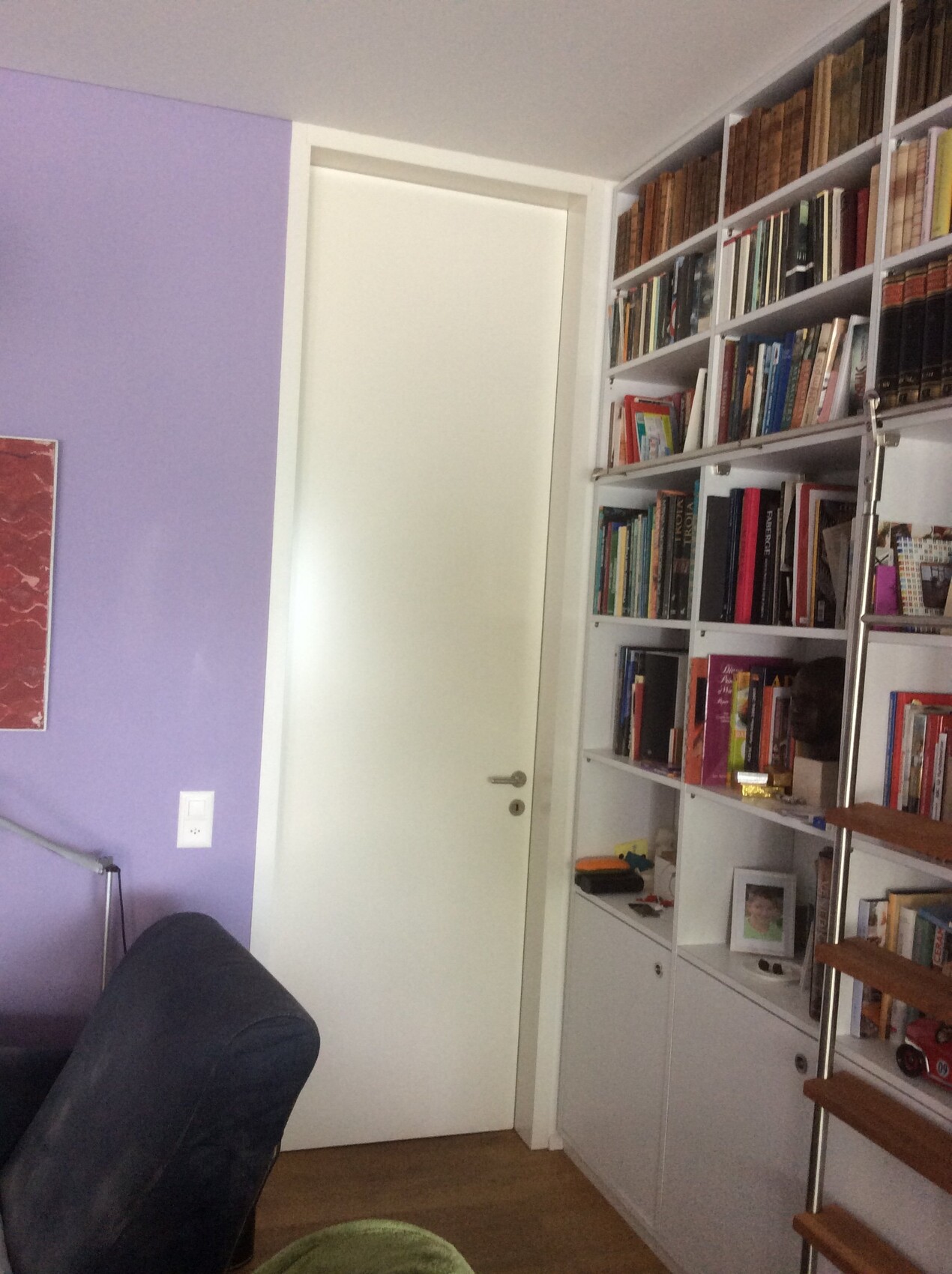 Bücherregal mit weißen Regalen, Tür in der Mitte, lila Wand und Leseecke.
