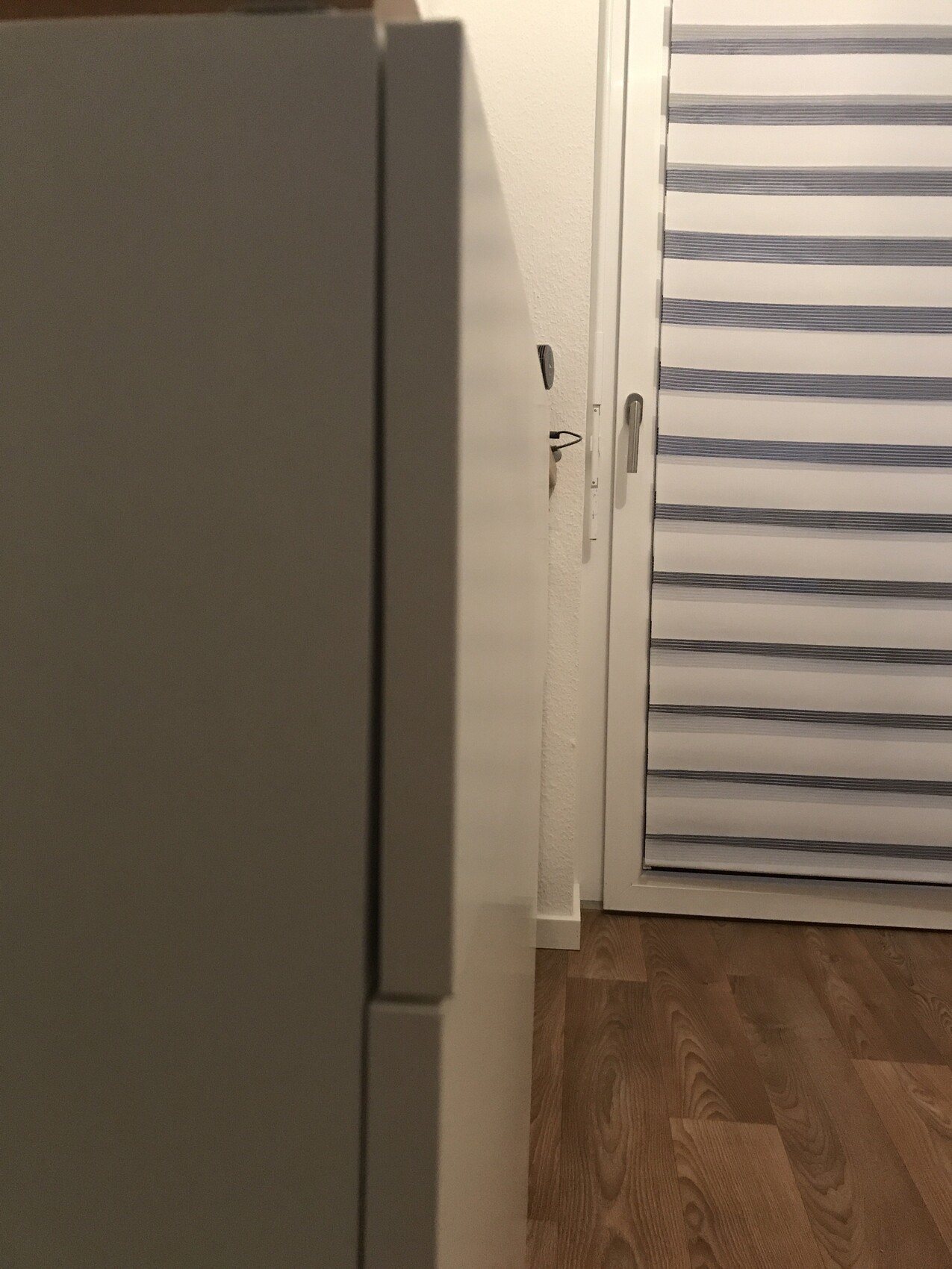 Innenraum-Ecke: grauer Schrank links, weiße Wand, horizontale Jalousie rechts, Holzboden.