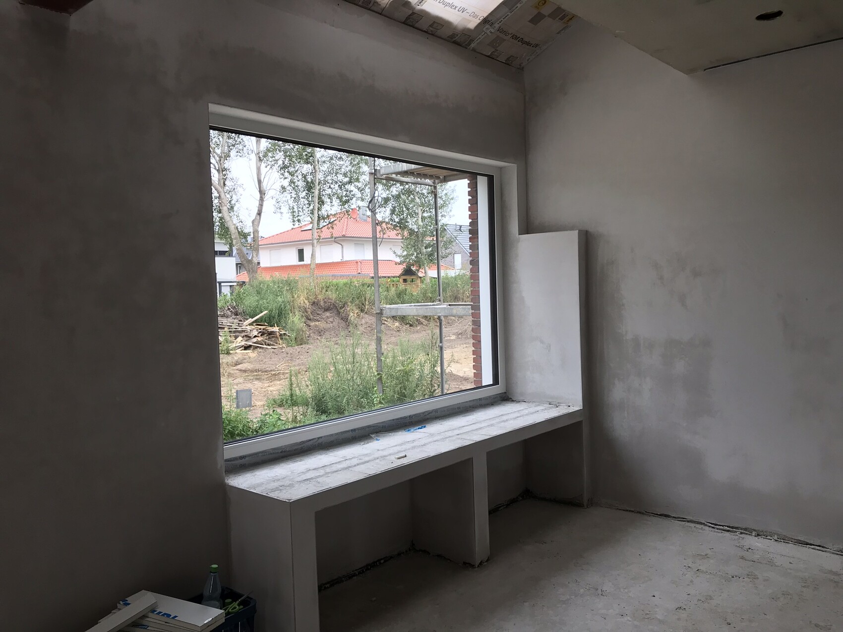 Rohbauzimmer mit großem Fenster, Blick auf Baugrube, Holzstapel und Häuser mit rotem Dach.