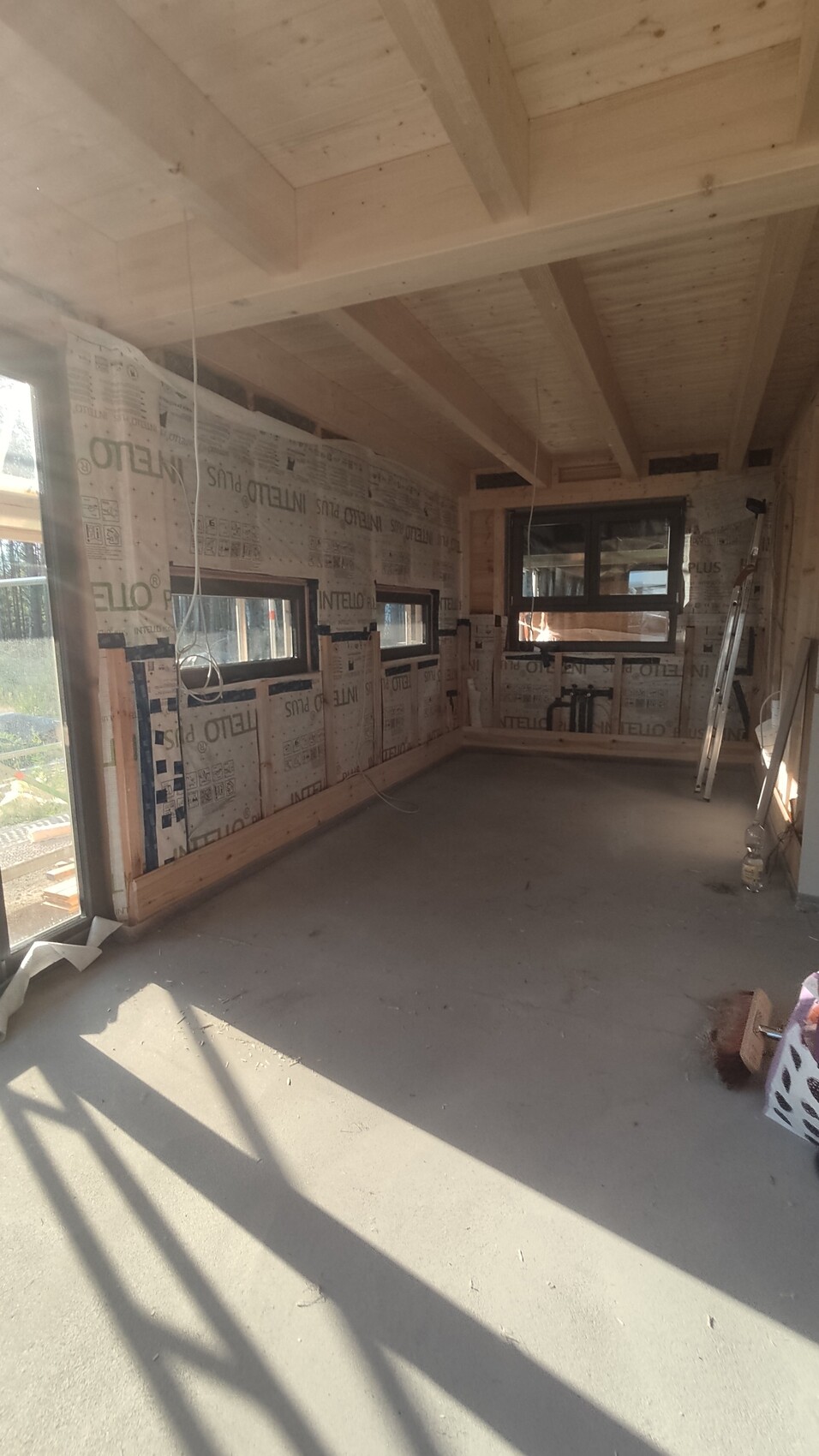 Unfertiger Innenraum im Bau mit Holzbalkendecke, Fenstern, Baufolie an den Wänden, Werkzeugen.