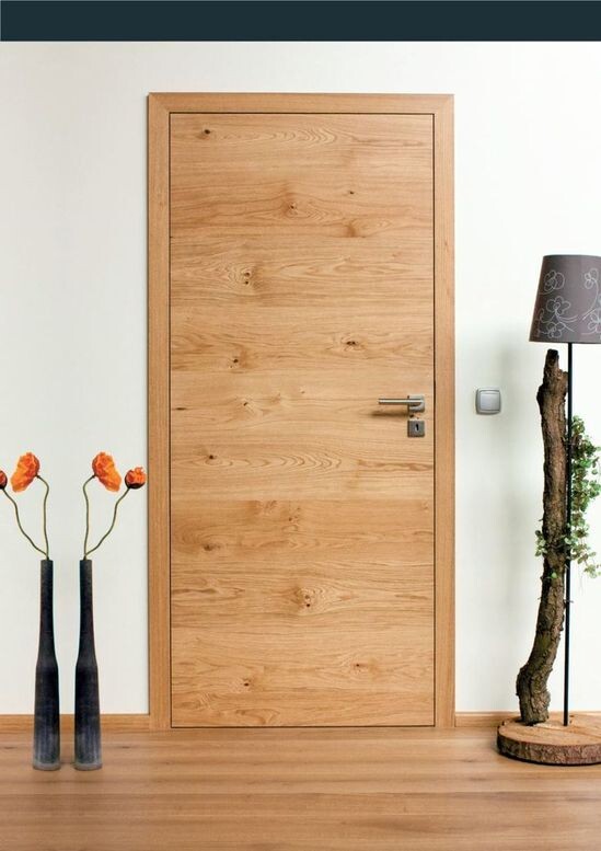 Heller Innenraum mit Tür aus hellem Holz; links Vasen mit orangefarbenen Blüten, rechts Stehlampe.