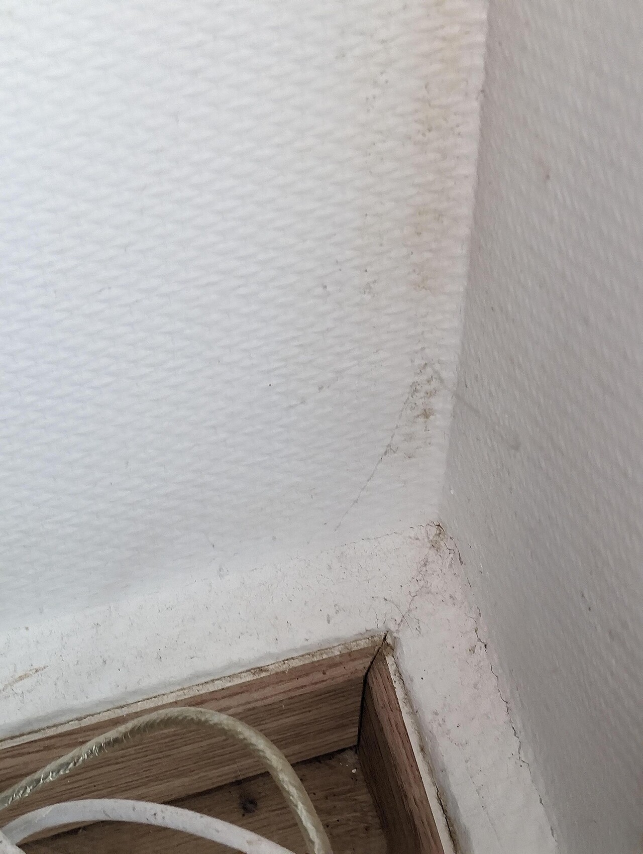 Eckbereich einer weißen Wand mit Fußleiste, Staubflecken und Riss; Kabel am Boden.