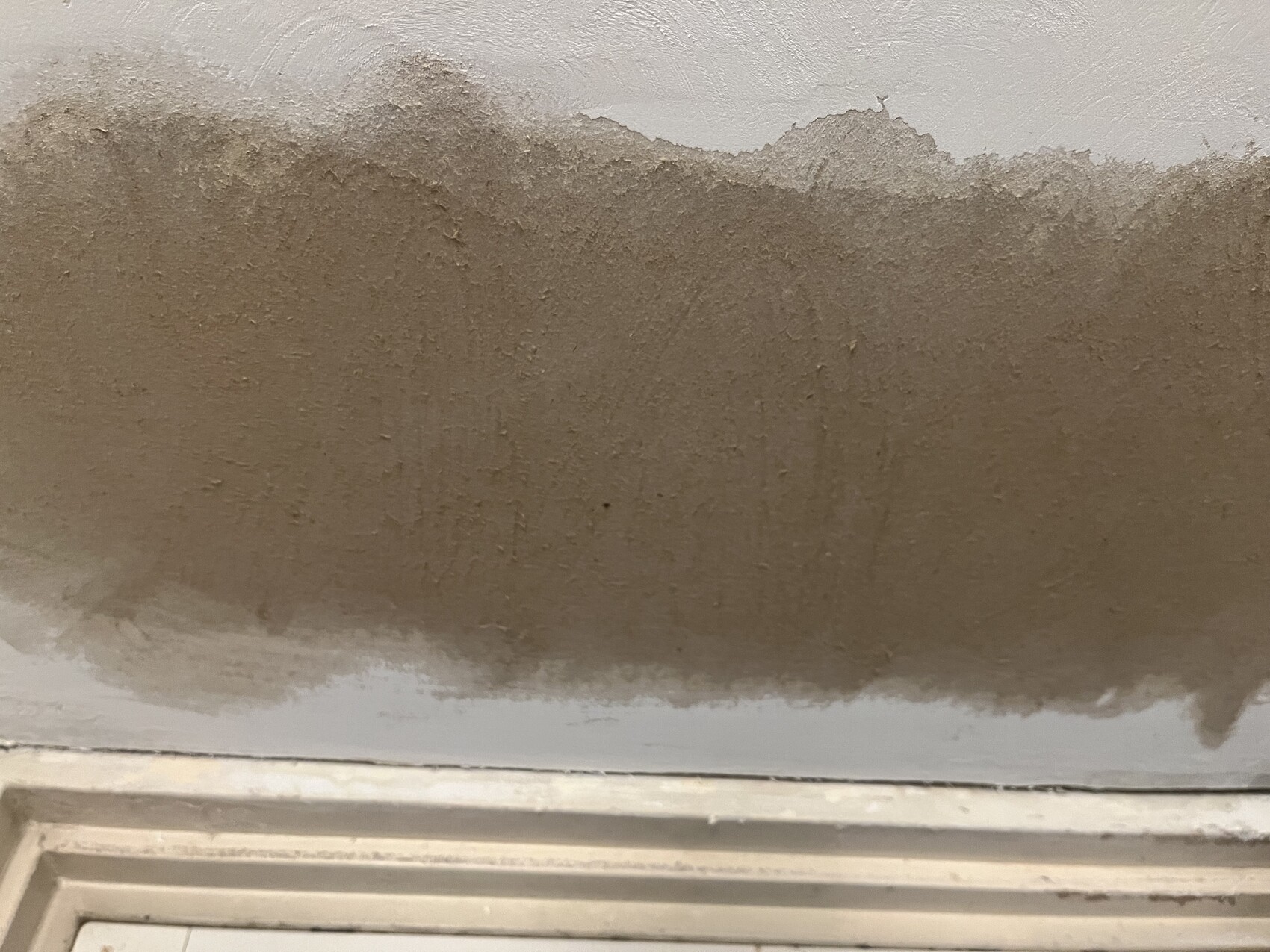 Braune, unregelmäßig strukturierte Putzwand mit rauer Oberfläche; unten weißer Fensterrahmen.