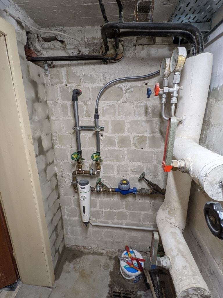Kellerraum mit Versorgungsrohren, Wasserfilter, Druckminderer, Zähler und Ventilen an der Betonwand.