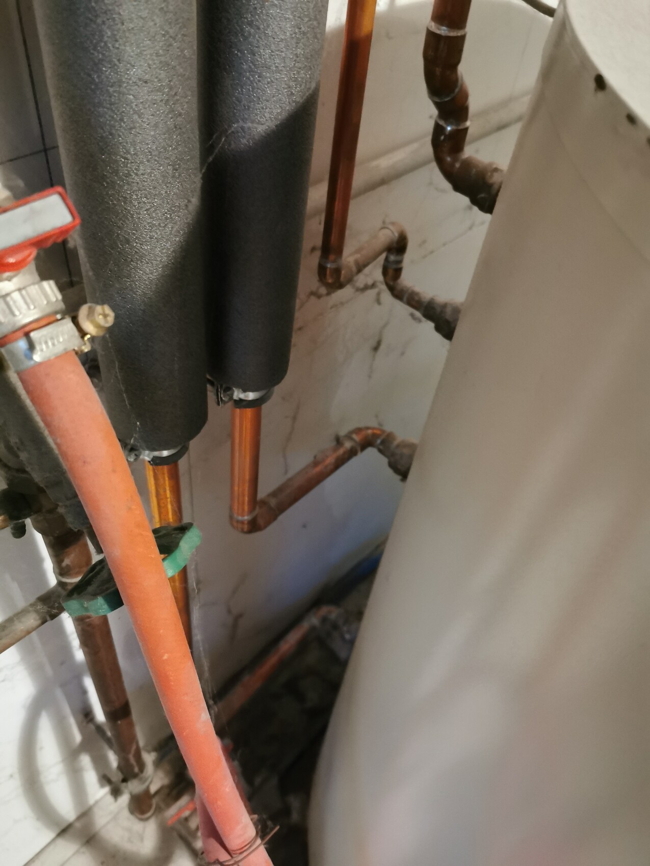 Kupferrohre mit schwarzer Dämmung, rotem Absperrventil und Warmwasserboiler im Keller.