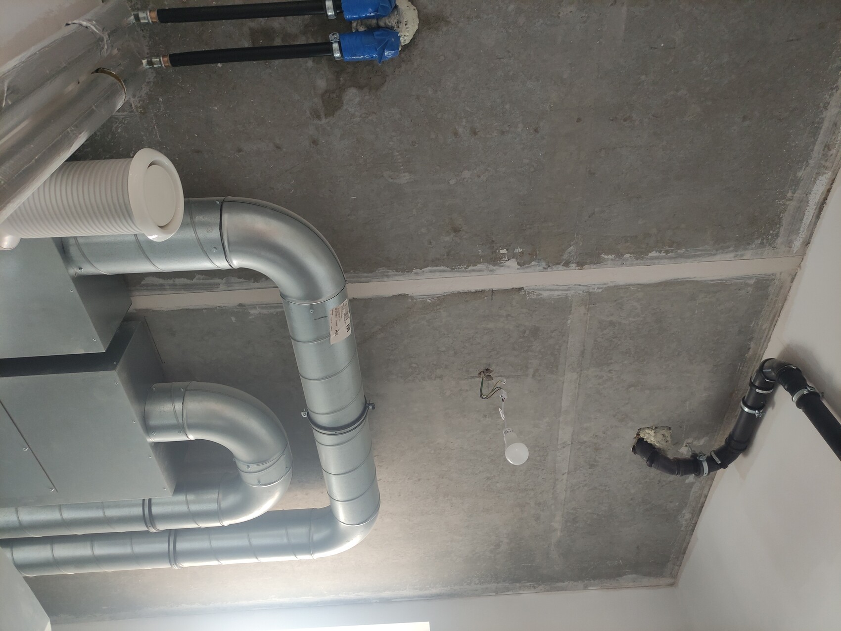 Exponierte Metallrohre und Lüftungsrohr ziehen über eine rohe Beton-Decke; lose Glühbirne hängt.