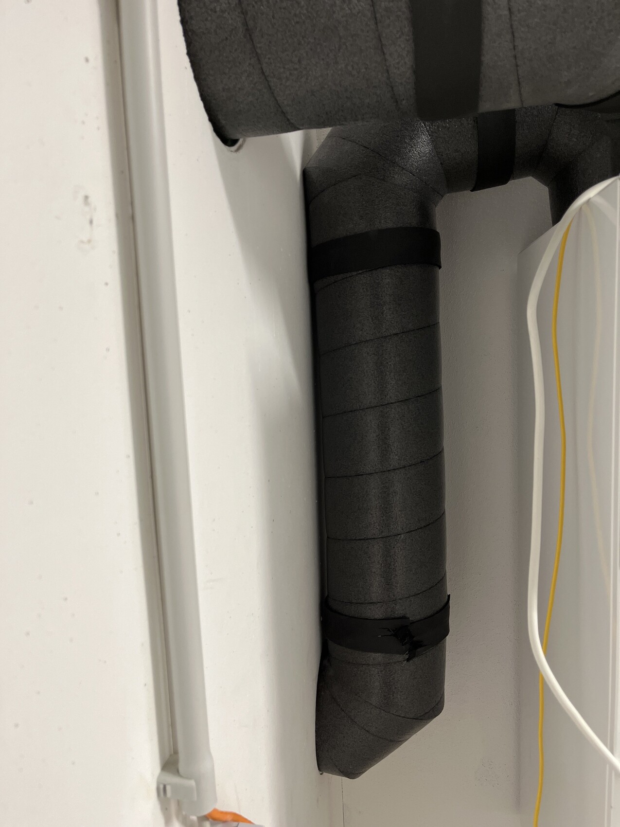 Schwarzer isolierter Schlauch mit Segmenten an der Wand; rechts weiße Box mit gelber Leitung.