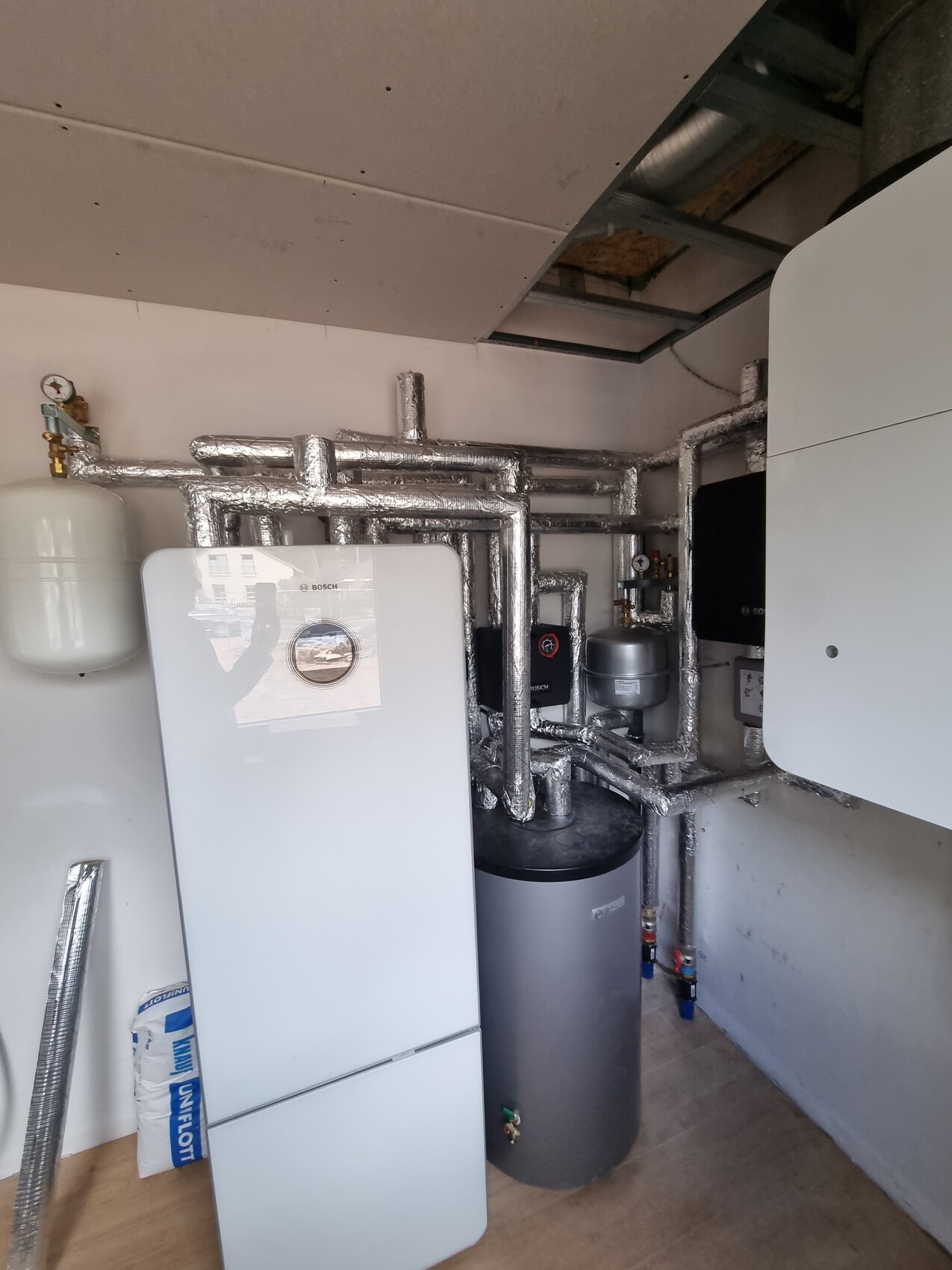 Heizungsraum mit weißem Boiler, stark isolierten Metallrohren, Ausdehnungsgefäß und Pumpe.