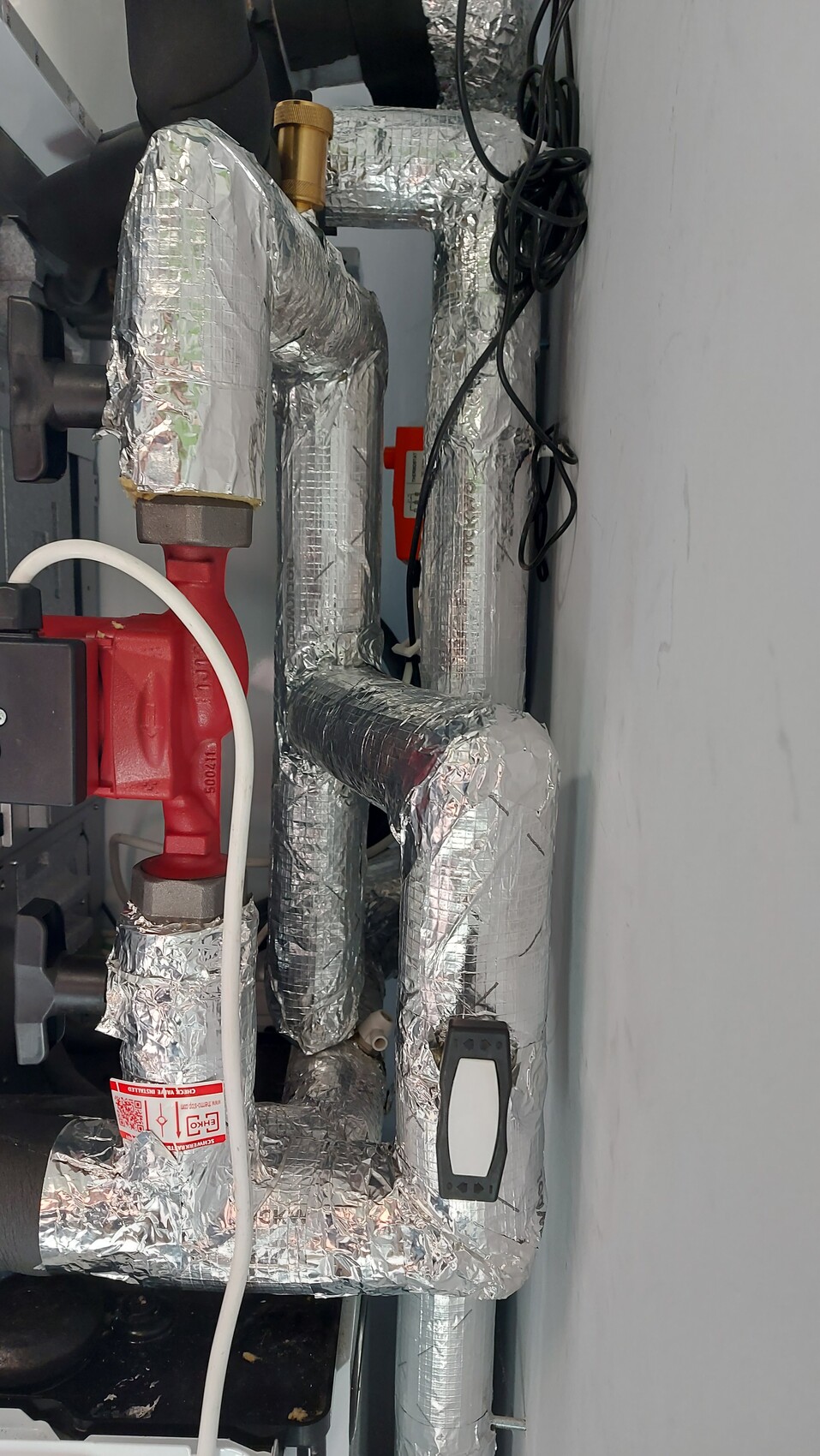 Isolierte Rohrleitungen in Alufolie, rotes Absperrventil und Kabelsalat im Technikraum.