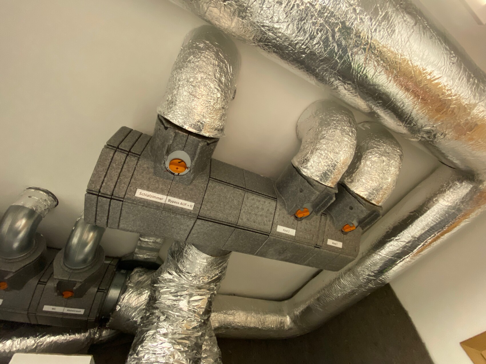 Isolierte Lüftungsrohre verbinden graue Verteilereinheiten mit orangefarbenen Ventilknöpfen.