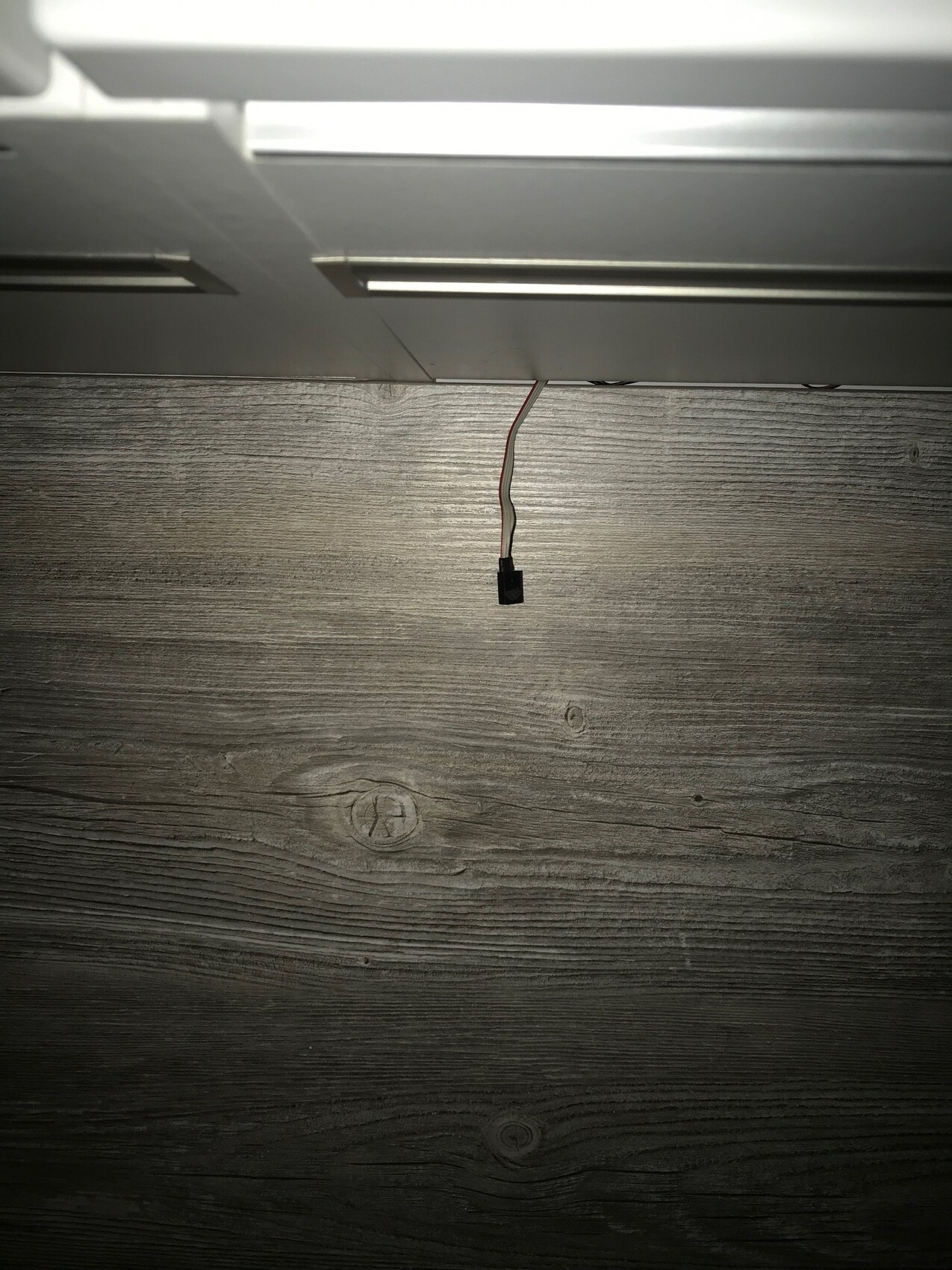 Hängendes Kabel mit schwarzem Steckverbinder über Holzfußboden unter weißem Möbelteil.