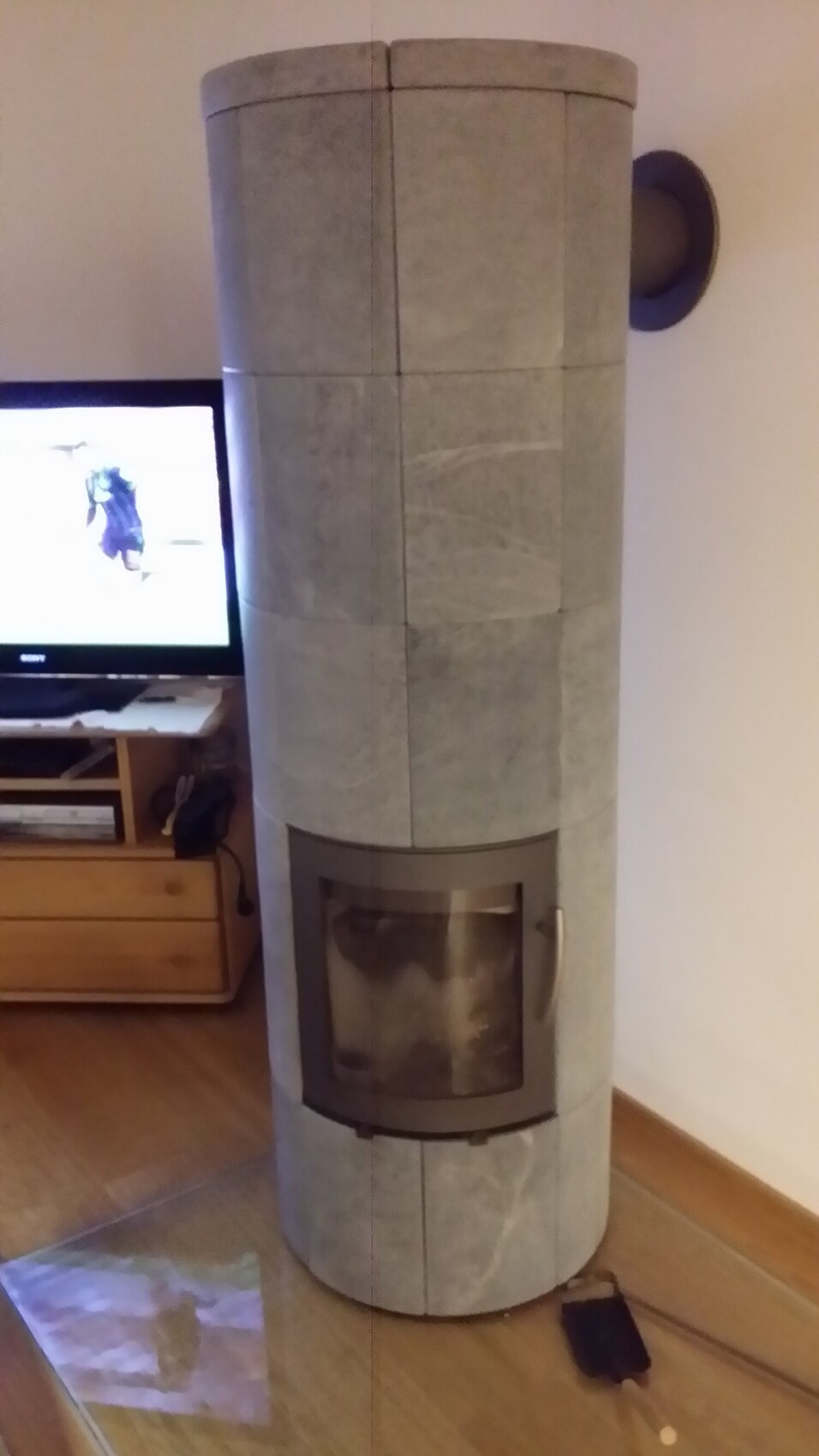 Großer runder Kaminofen aus Stein mit Glasfront im Wohnzimmer neben dem Fernseher.
