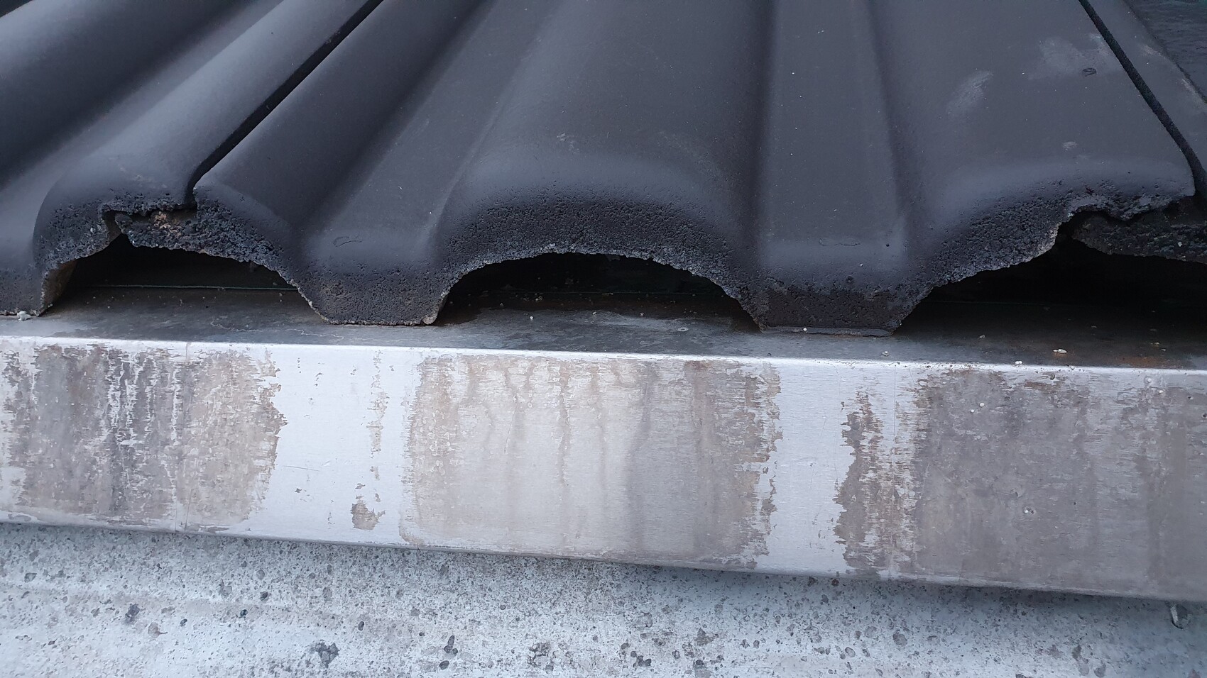 Nahaufnahme dunkler Dachziegel mit abgebrochener Kante; darunter verschmutzte Metallverkleidung.