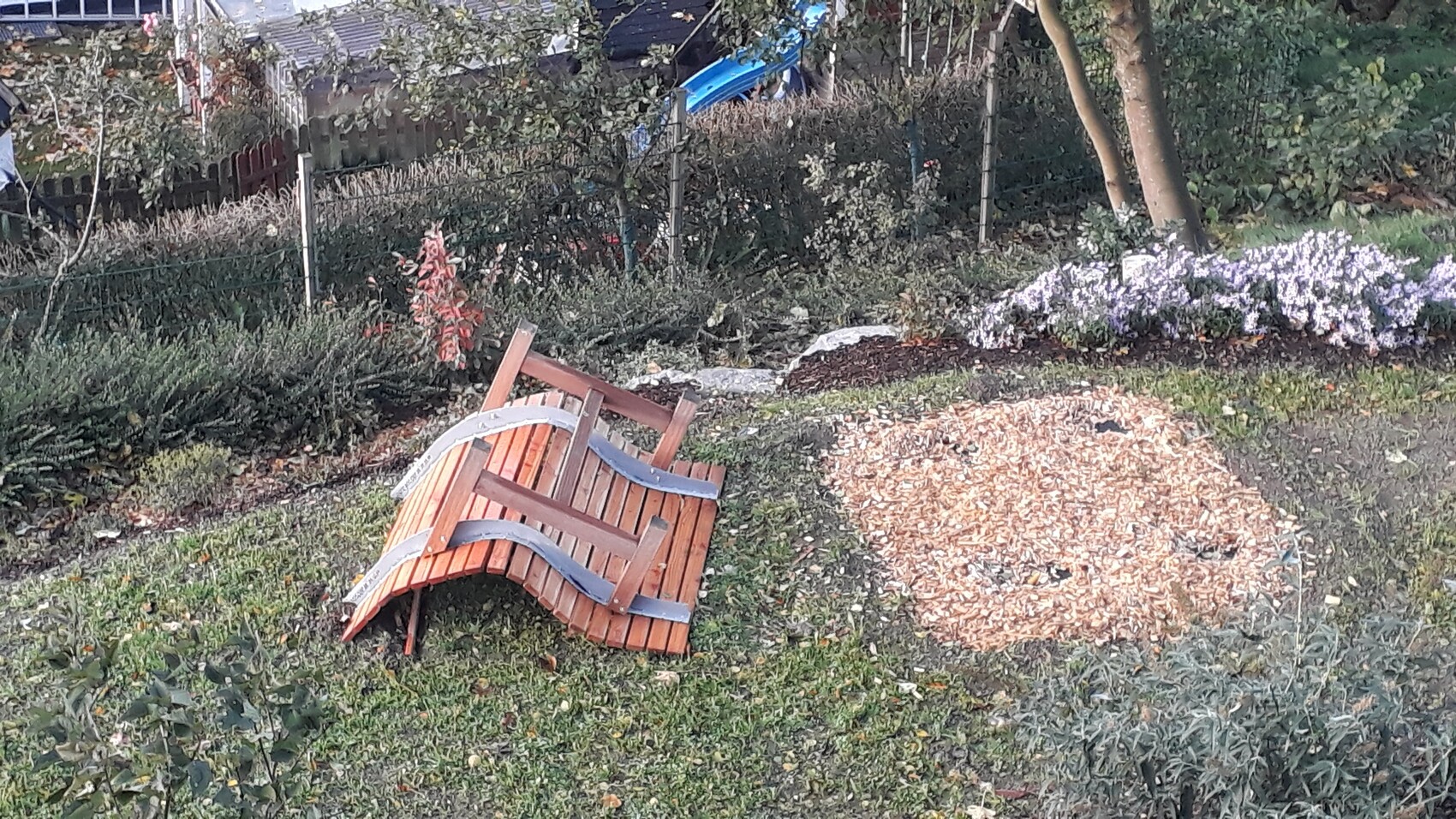 Umgefallene Garten-Holzbank mit Metallstreben neben Holzschnitzel-Haufen; Hintergrund mit Sträuchern und Zaun.