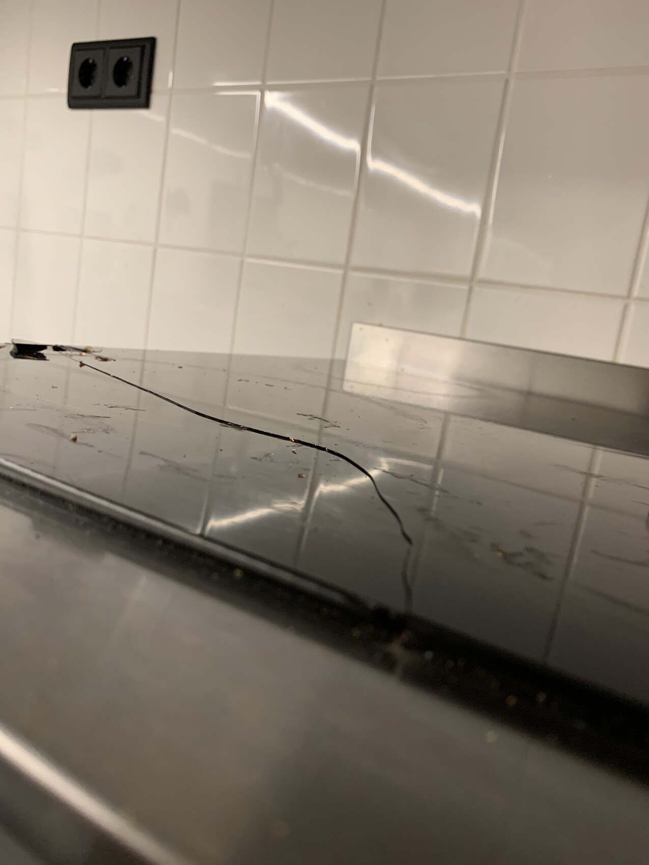 Schwarzes Glaskeramik-Kochfeld mit Haarriss, Spiegelung der weißen Fliesenwand im Hintergrund.