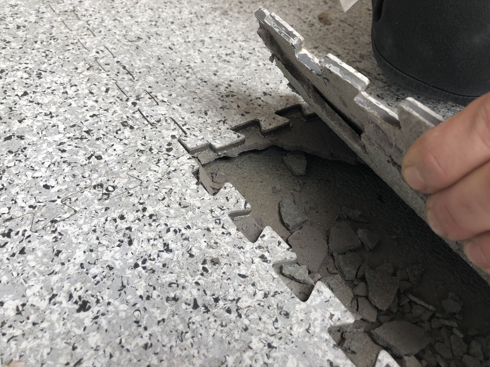 Hand hebt eine zerbrochene Fliese an; darunter dunkler Zementboden mit Bruchstücken.