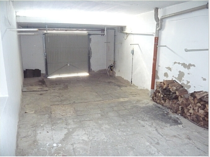 Leerer Keller-/Garagenraum mit offenem Tor am Ende, Holzstapel rechts.