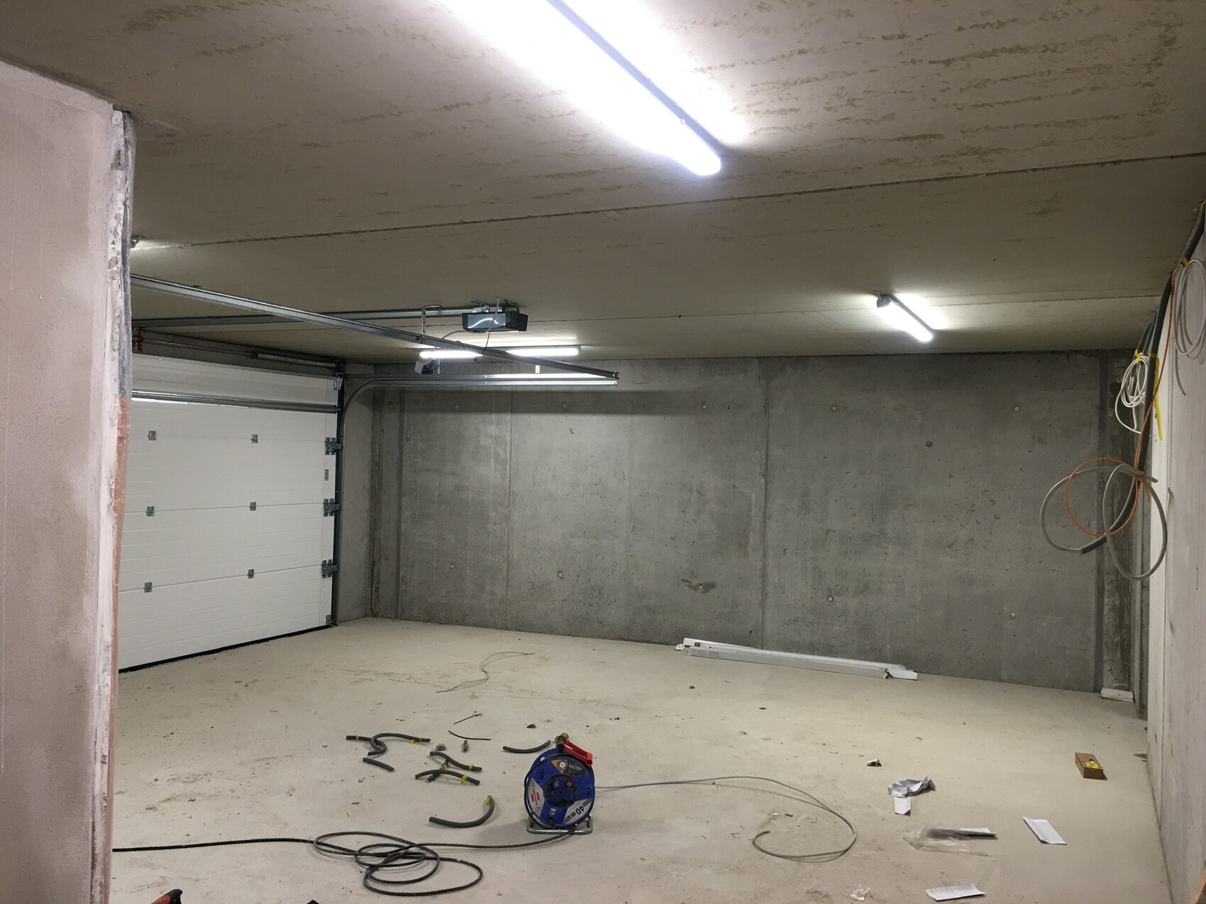 Leere Beton-Garage mit weißem Rolltor, Kabeln an der Wand und Werkzeugen auf dem Boden.