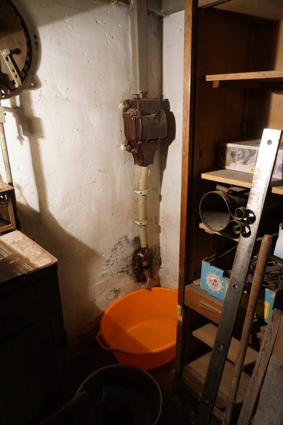 Kellerraum mit braunem Verteilerkasten an Rohr, orange Kunststoffwanne, Regal mit Werkzeug.