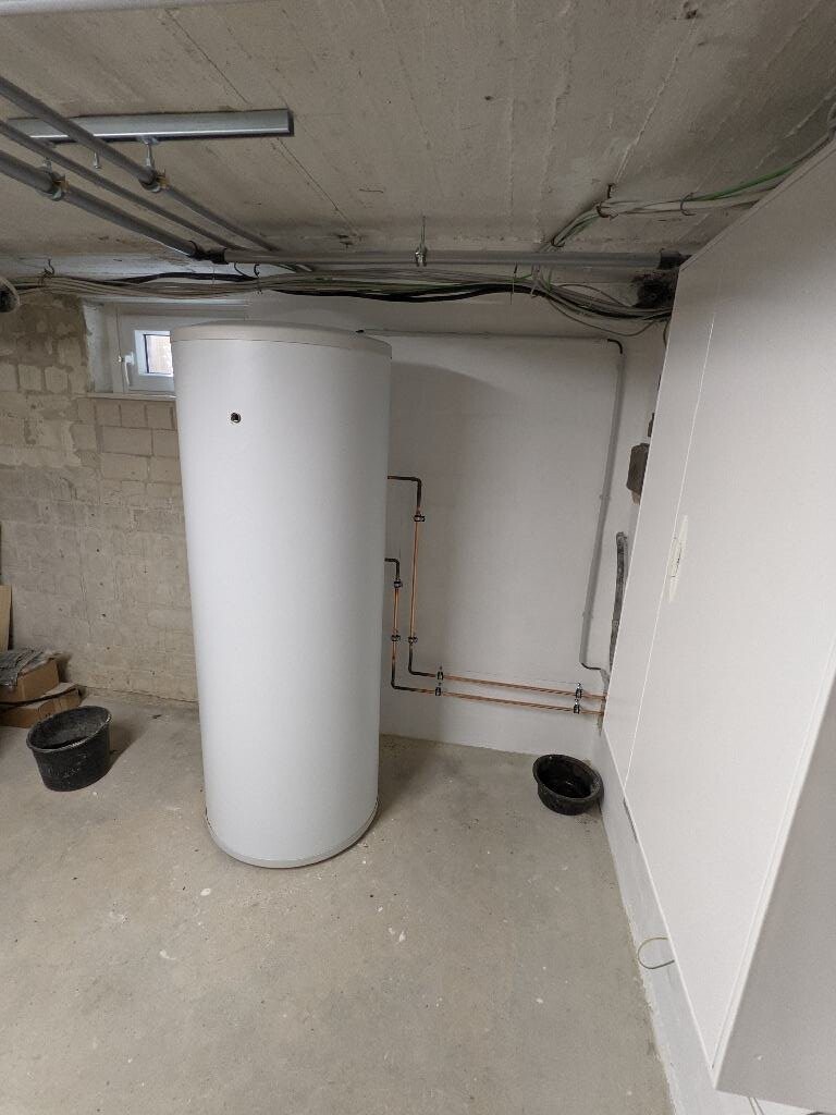 Großer weißer Warmwasserspeicher im Keller, Rohre an der Wand, Eimer daneben.