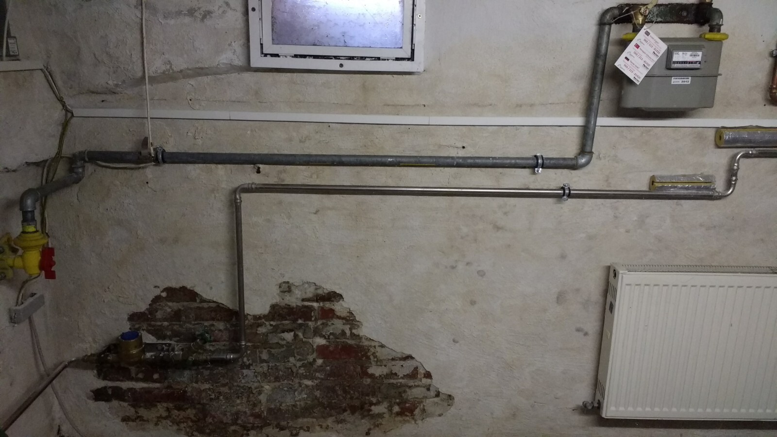 Kellerwand mit sichtbaren Rohren, Gasventil, Heizkörper und kleinem Fenster