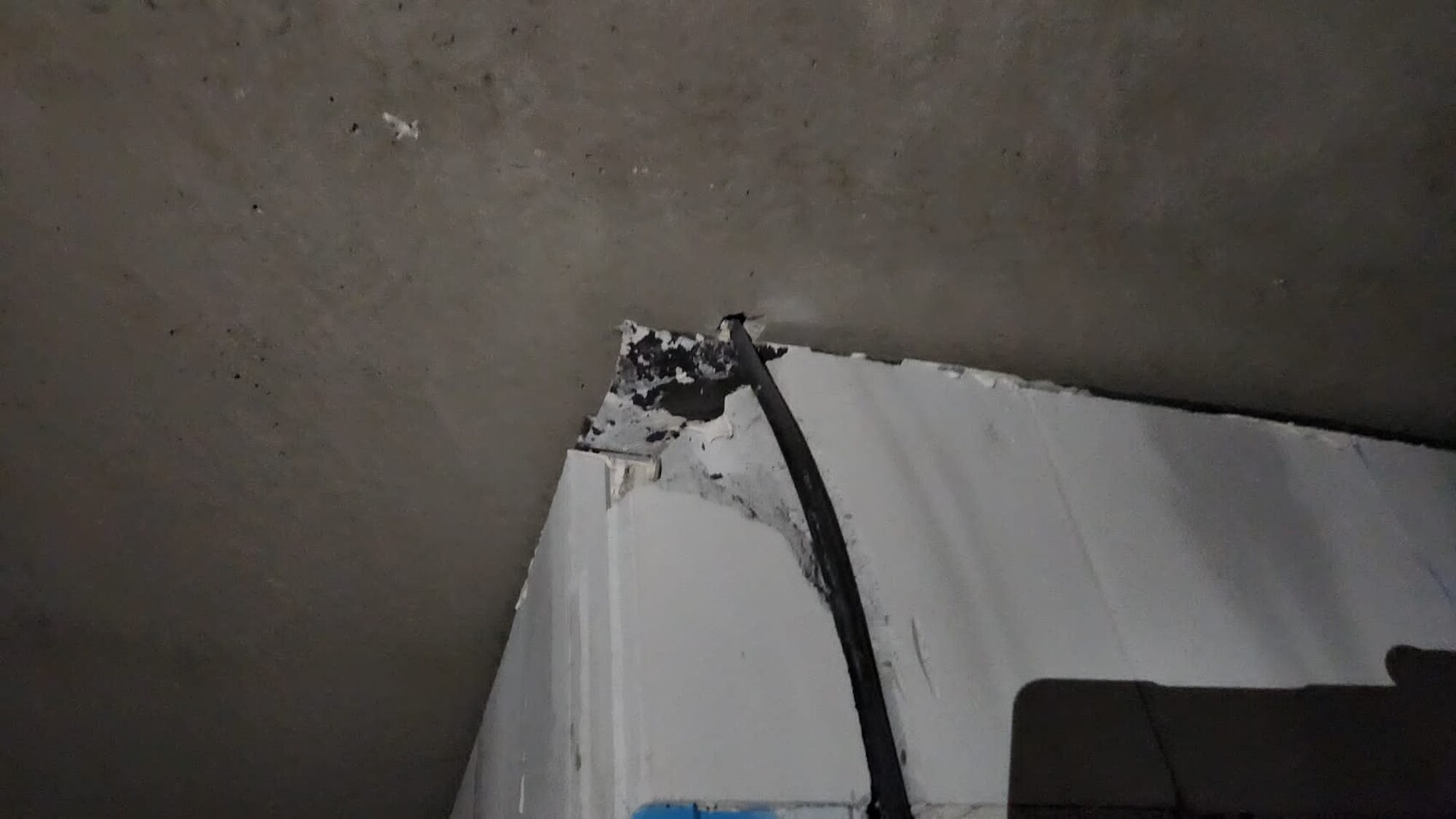 Schwarzes Kabel verläuft entlang einer brüchigen, weiß gestrichenen Wandkante in einem dunklen Raum.
