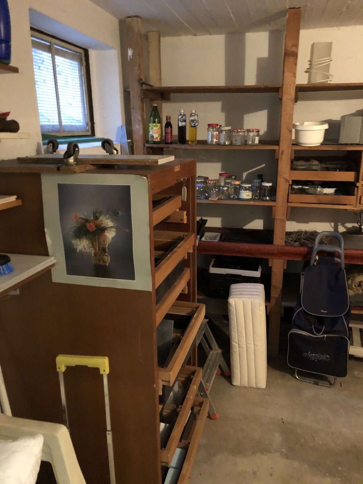 Keller/Werkstatt mit Holzregalen, Gläsern, Flaschen, Poster am Schrank und blaue Tasche.