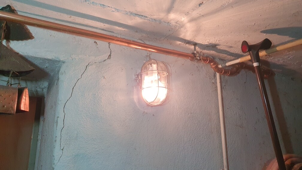 Kellerraum: blaue Wand mit Rissen, Schutzgitter-Lampe an der Decke, Rohrleitungen.