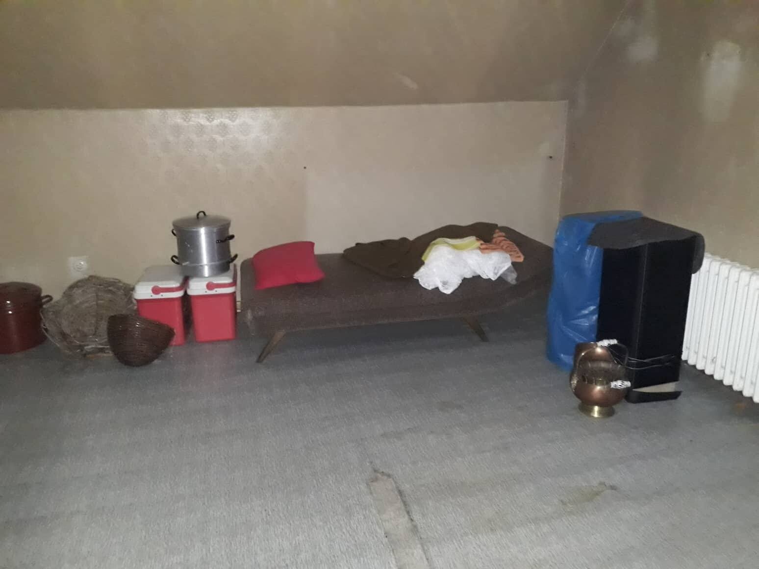 Kellerraum mit Bett, Decke, Kleidung, zwei roten Kühlboxen mit Topf darauf, Körbe.