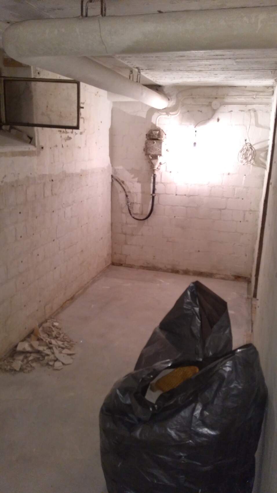 Kellerraum mit weißen Ziegelwänden, sichtbaren Rohren, Elektrokasten und schwarzem Müllsack.