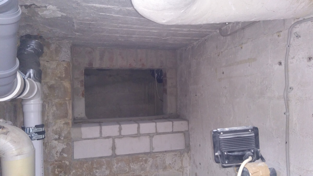 Kellerraum mit rechteckiger Backstein-Nische in der Wand; Rohre links, Kabel rechts.