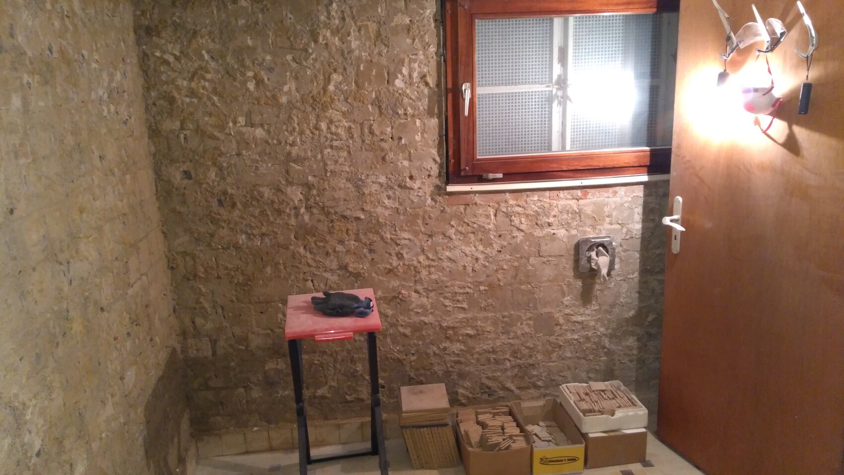 Kellerraum mit rauer Steinwand, Fenster, Holzrahmen; roter Tisch mit Handschuhen, Kisten mit Fliesen.
