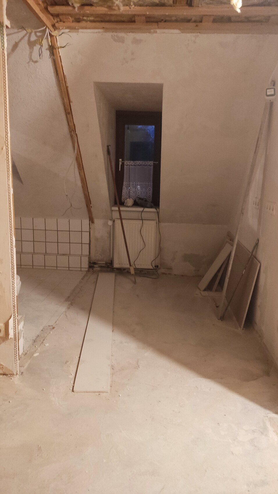 Unrenovierter Dachboden mit schräger Decke, Holzbalken, Fenster mit Spitzenvorhang und Heizkörper.
