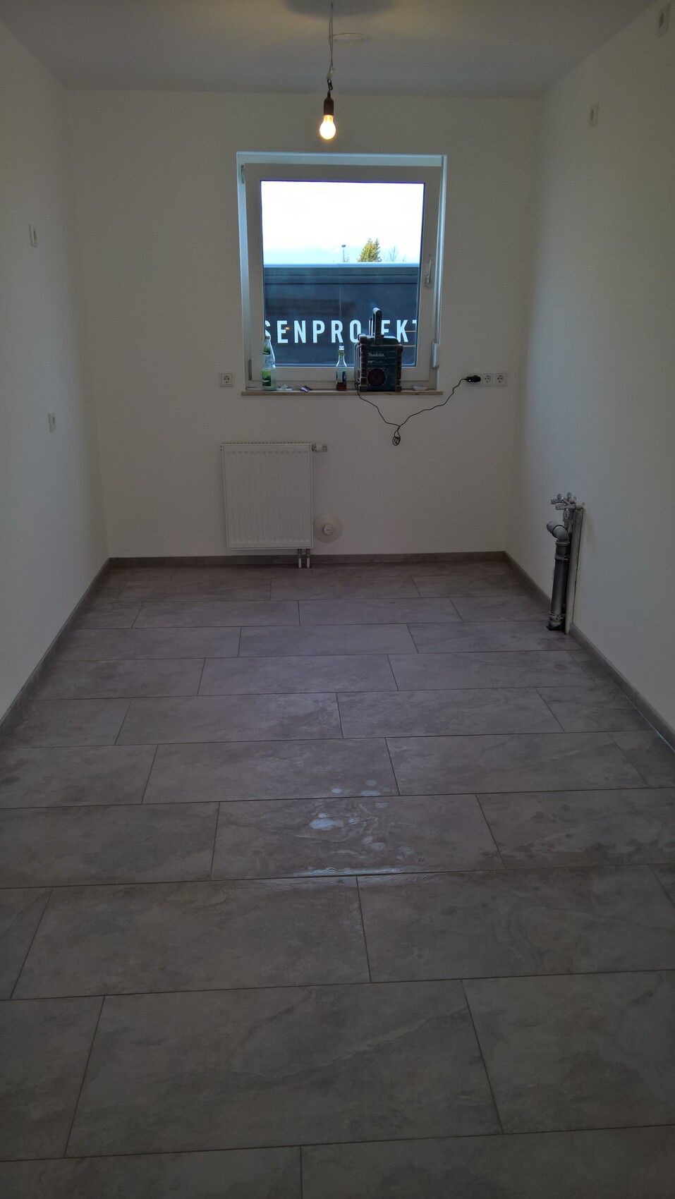 Helles, leeres Zimmer mit Fliesenboden, hängende Glühbirne, Fenster, Heizkörper und Rohrleitungen.