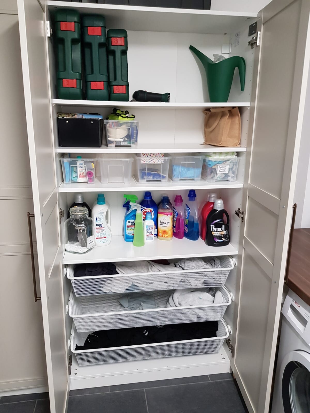 Offener Schrank mit Reinigungsmitteln, kleinen Aufbewahrungsboxen, Werkzeugkisten und Wäschekörben.