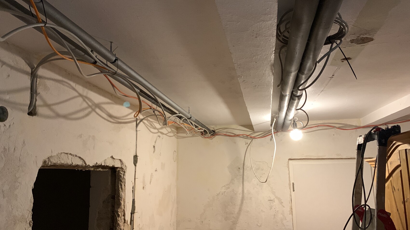 Kellerraum mit freiliegenden Kabeln, Rohren an der Decke und einer hängenden Glühbirne.