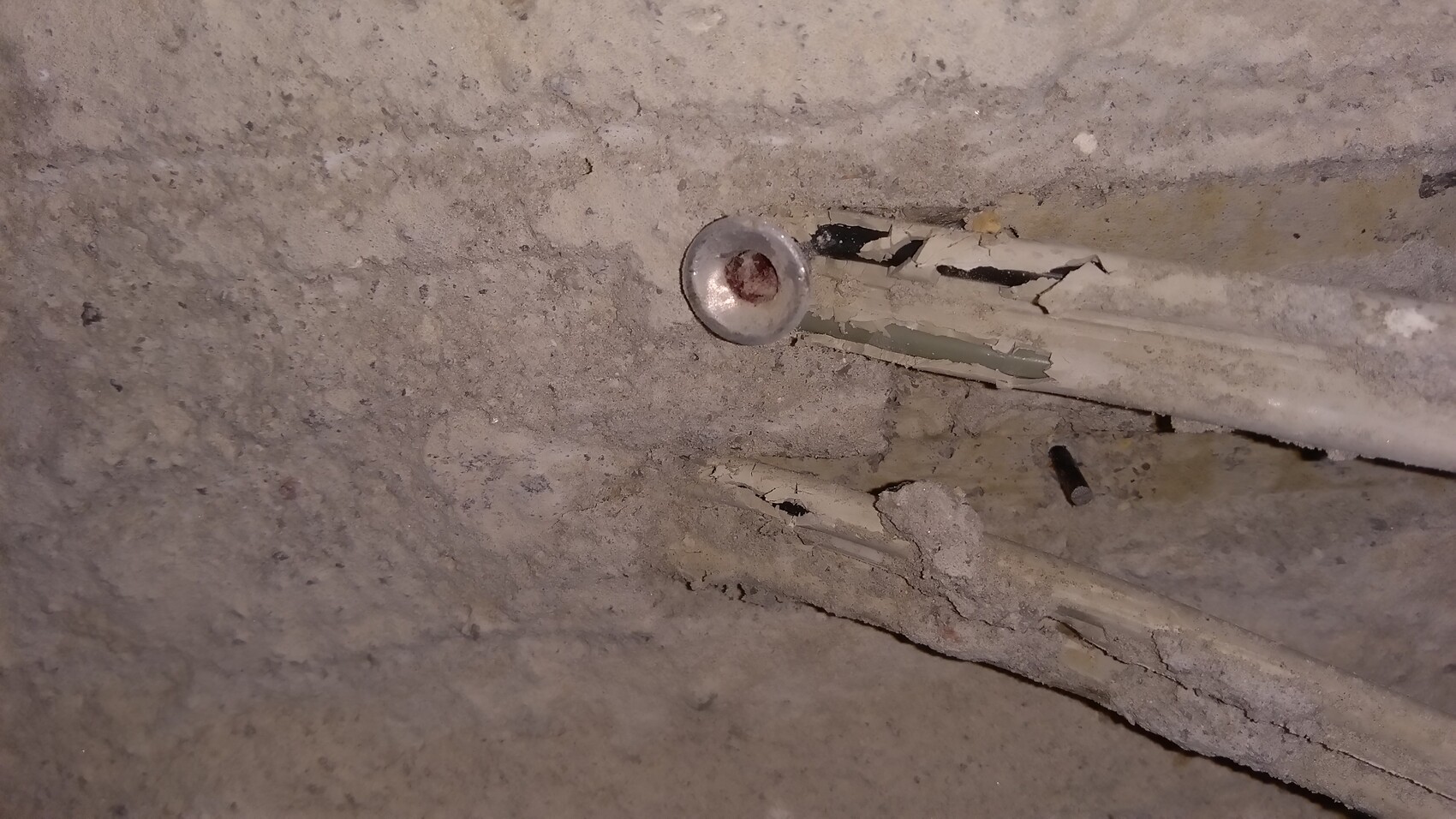 Runde Metallkappe in staubiger Betonwand, daneben bröckelige Leitung und Risse.