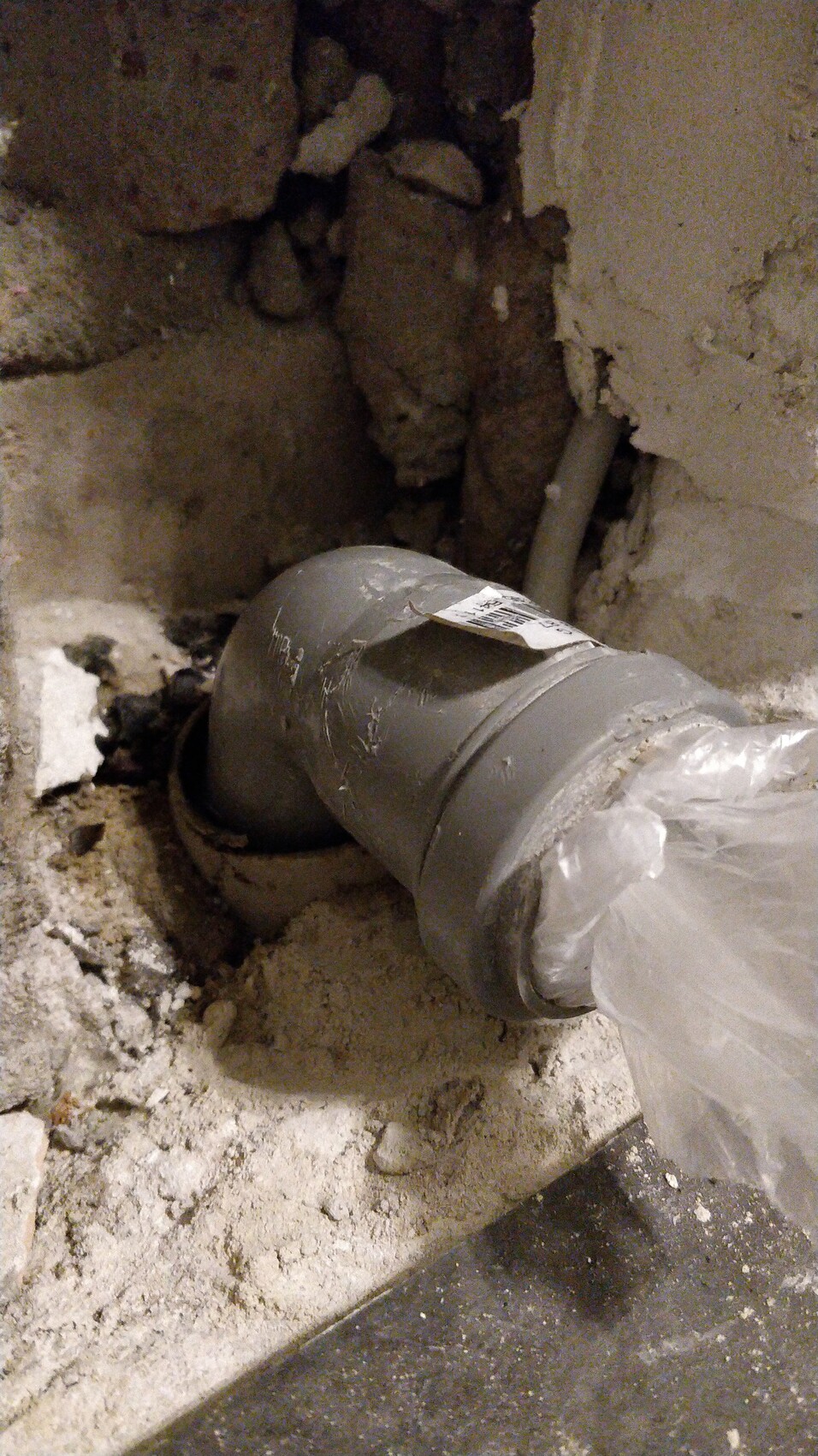 Grau Abflussrohrbogen in staubigem Keller, beschädigte Wand, Plastikfolie am Ende.