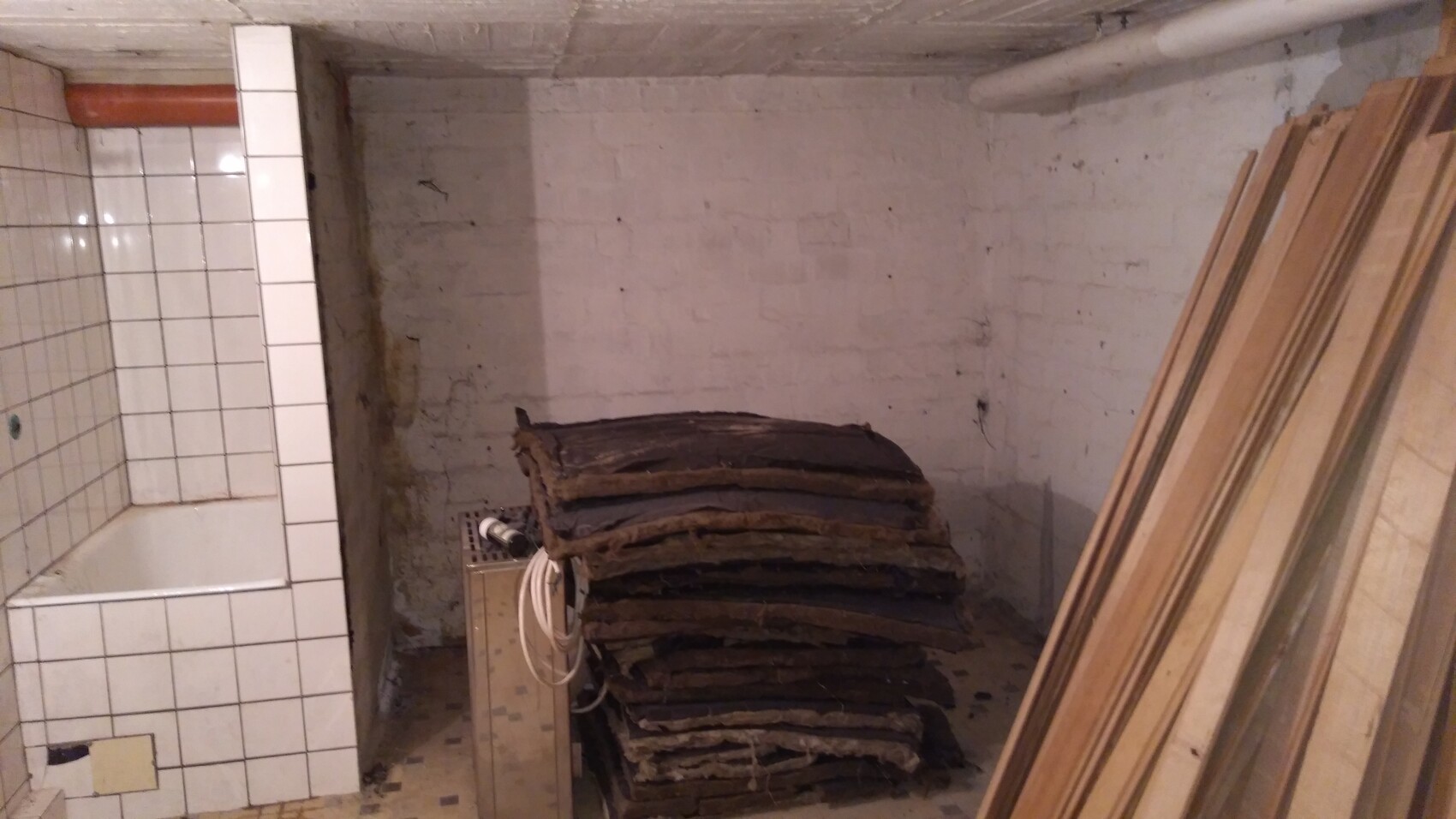 Dunkler Kellerraum mit Stapel alter Dämmplatten, rechts Holzleisten, links Badewanne.