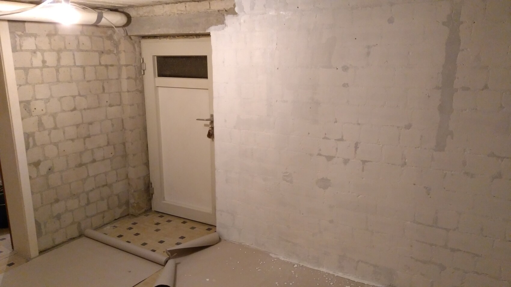 Unrenovierter Kellerraum: weiße Ziegelwände, Tür mit kleinem Fenster, Bodenrollen liegen da.
