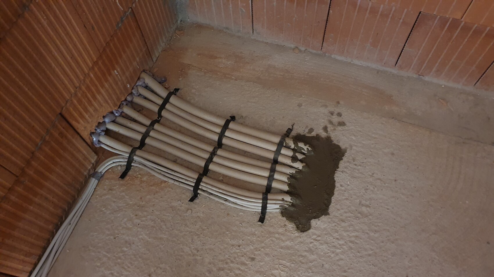 Mehrere weiße Kabelrohre liegen gebündelt am Boden in der Ecke der Kellerwand, daneben Zementschlick.