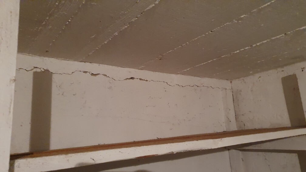 Kellerraum: weiße Wand mit Rissen, darunter ein Holzregal.