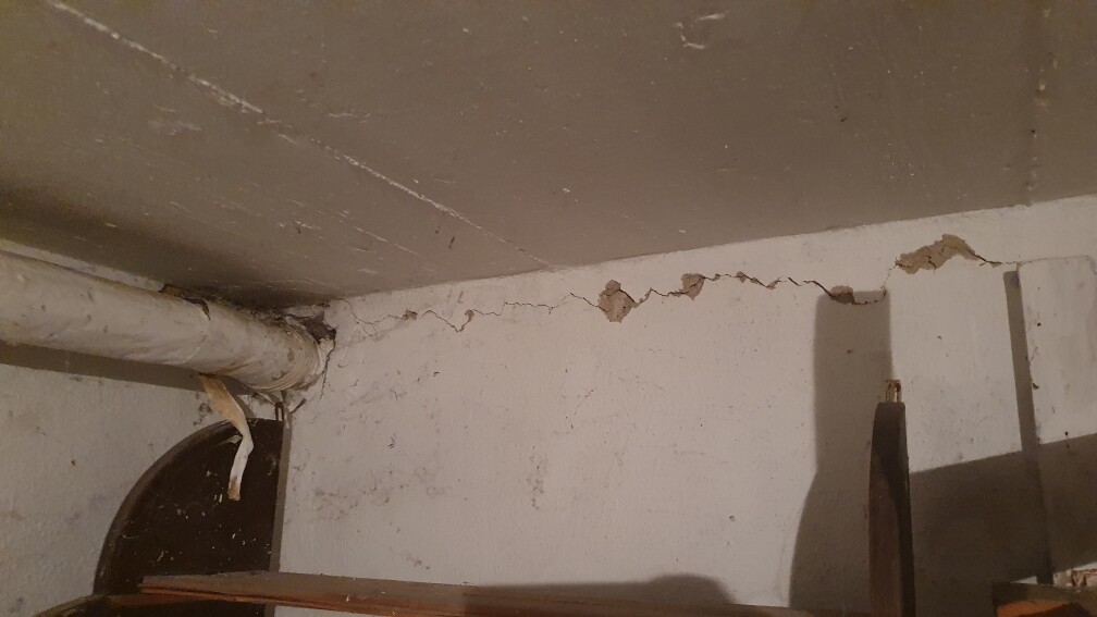 Kellerwand mit Rissen, abplatztem Putz und einem Rohr an der Decke.