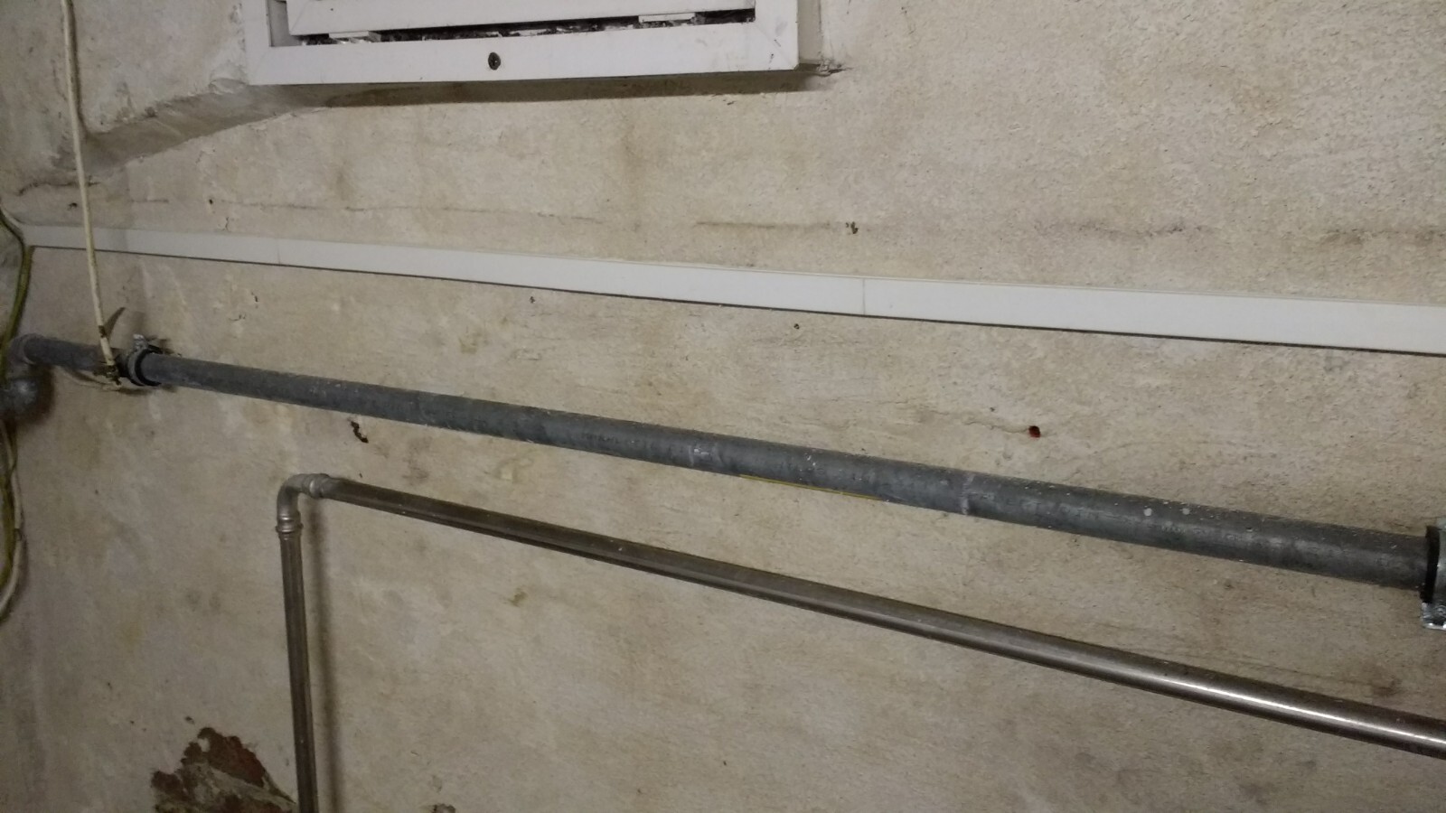 Kellerwand mit sichtbaren Metallrohren, rauer Putz und kleinem Fenster
