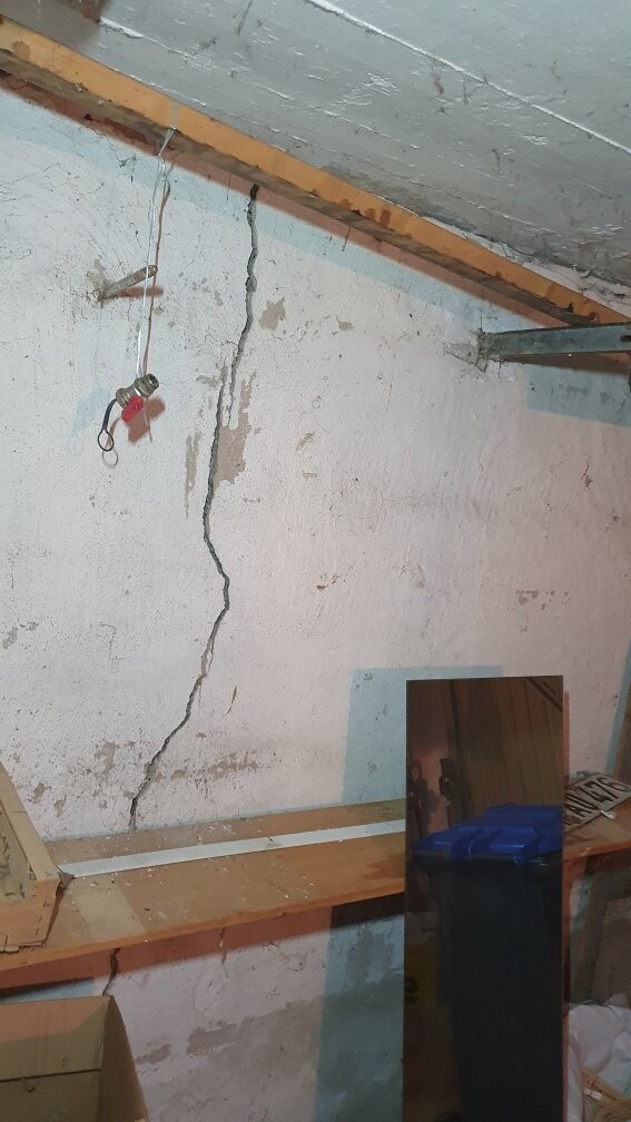 Großer Wandriss im Keller, lose Kabel, hängende Lampe, Spiegel und Holzregal.