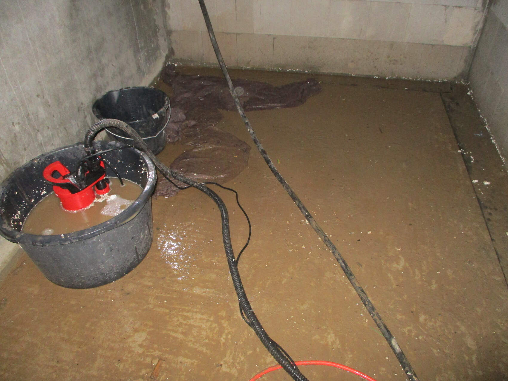 Keller mit stehenden braunen Wasserpfützen, Eimer mit Pumpe und Schlauch auf nassem Boden.